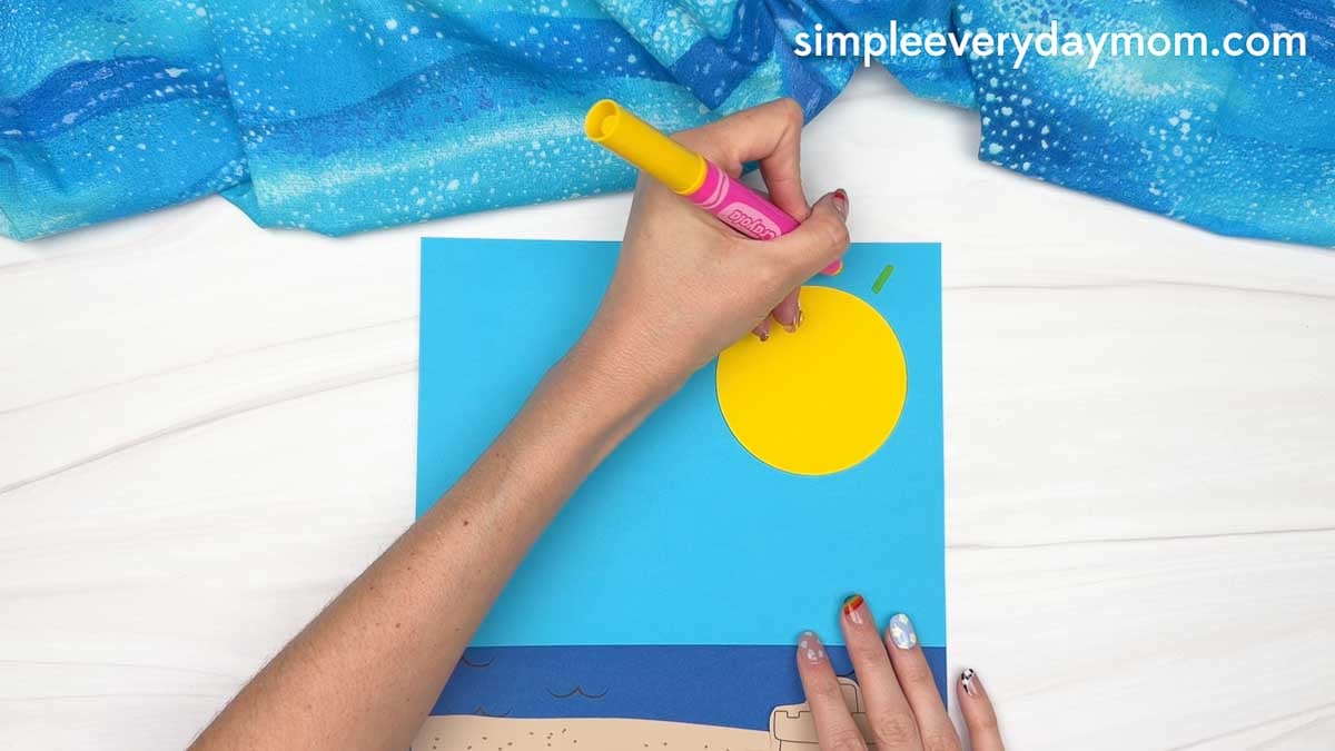 Hands draw sun rays around a yellow paper sun on a beach scene background for a Christmas in July gingerbread man craft.