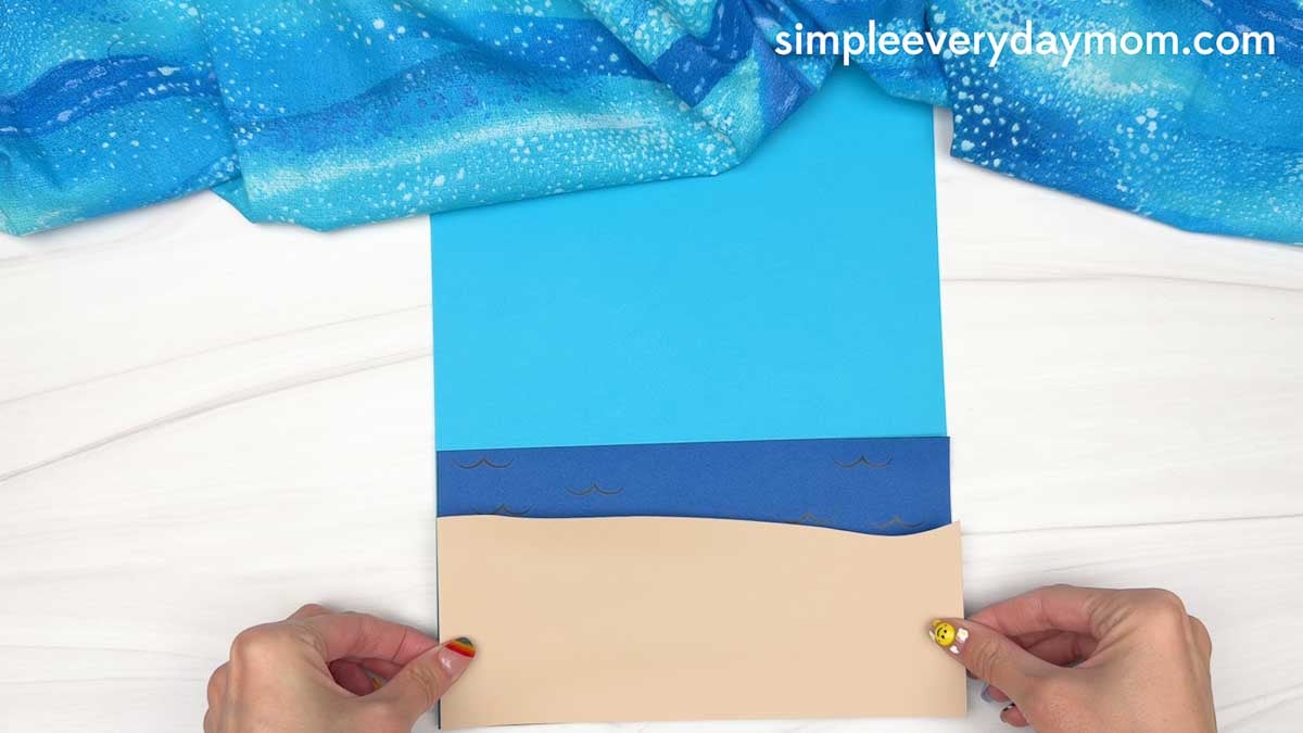 Hands position a tan cardstock piece on a blue ocean and sky background for a Christmas in July gingerbread man beach craft.