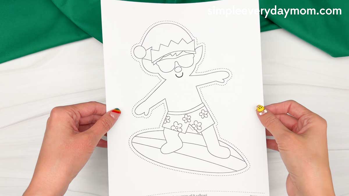 Hands holding a printed elf surfing craft template with a beach elf wearing sunglasses, shorts, and a Santa hat standing on a surfboard.