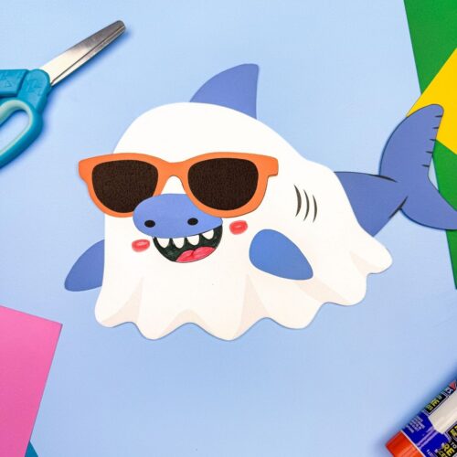 A completed craft of a Summerween ghost shark wearing orange sunglasses on a blue background with craft supplies.