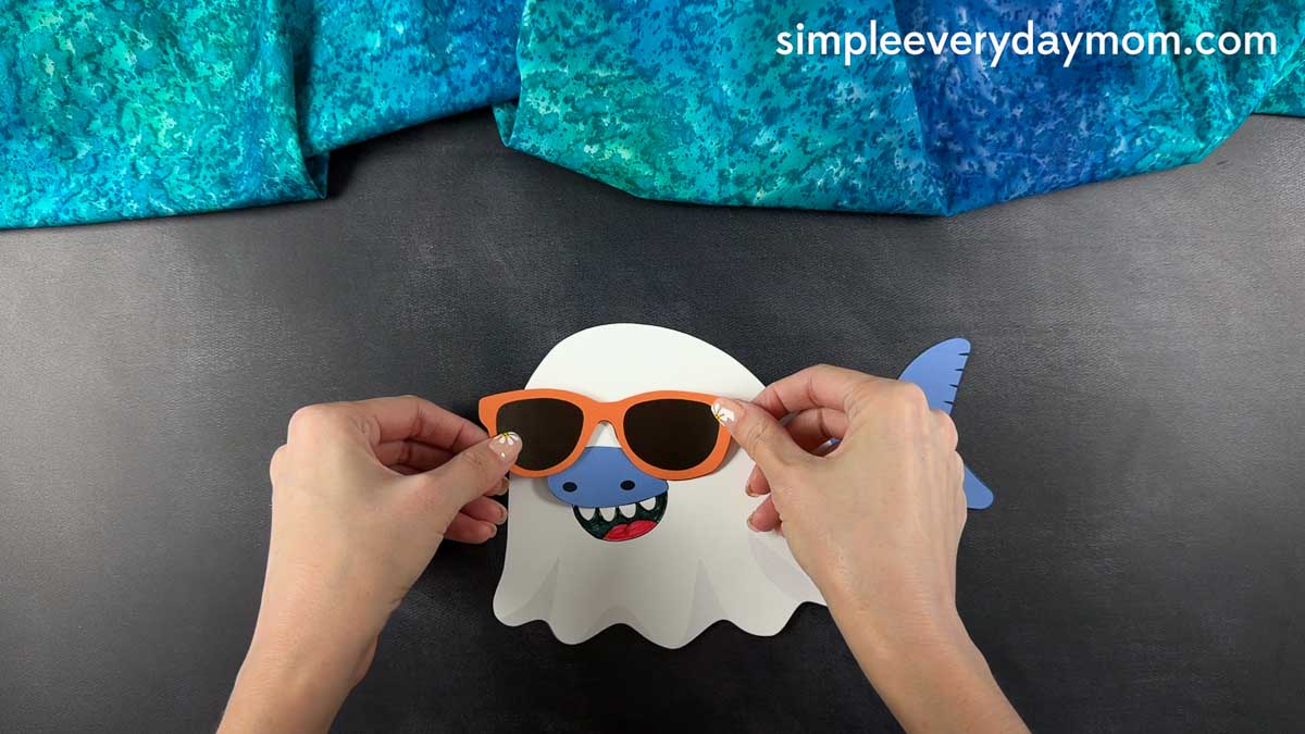 Hands placing orange sunglasses onto a partially assembled Summerween ghost shark craft.