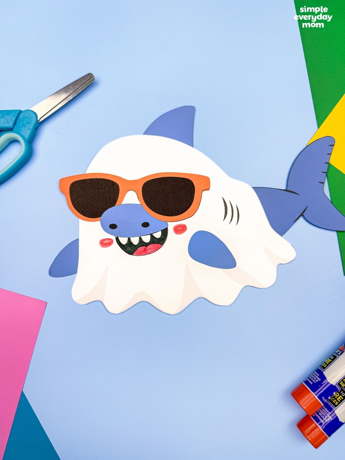 A completed craft of a Summerween ghost shark wearing orange sunglasses on a blue background with craft supplies.