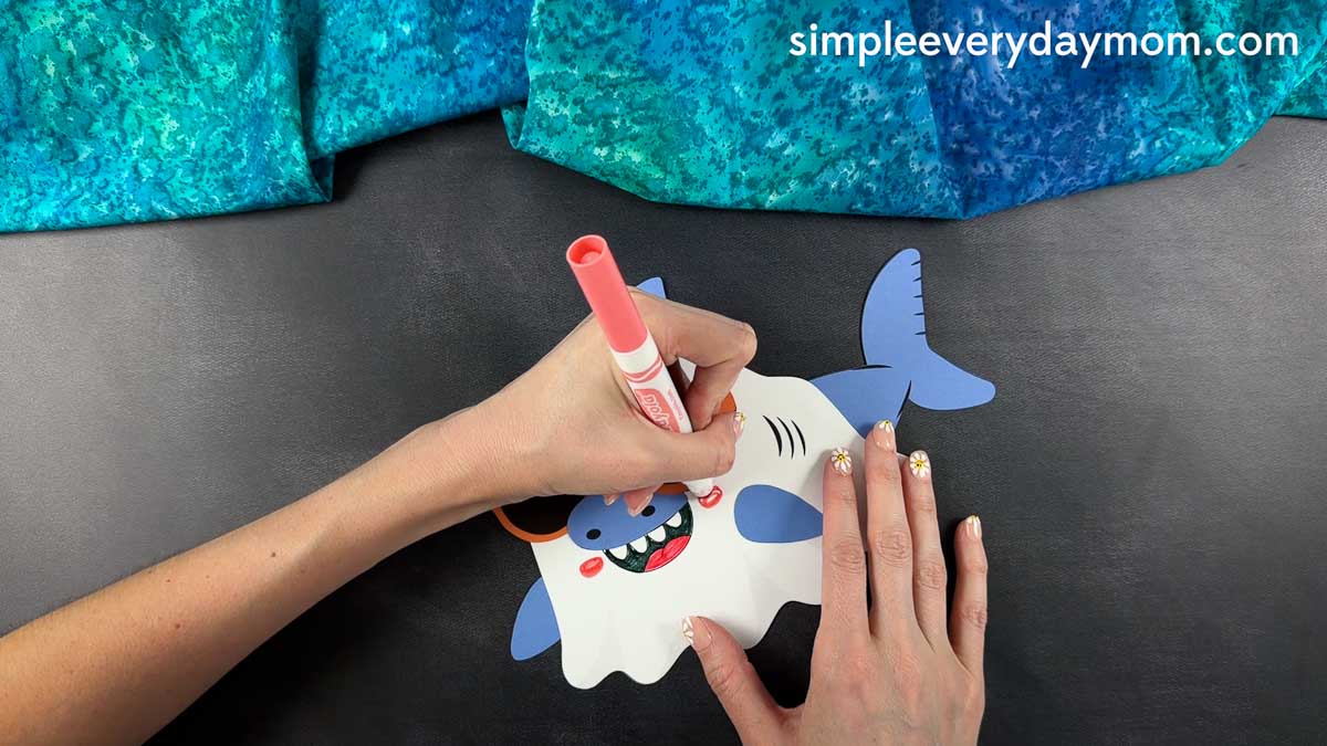 Hands drawing rosy cheeks on a nearly finished Summerween ghost shark craft with a pink marker.