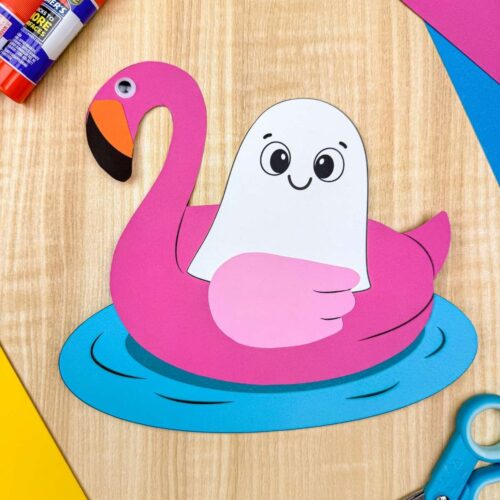A completed craft featuring a Summerween ghost floating in a pink flamingo pool float surrounded by craft supplies.