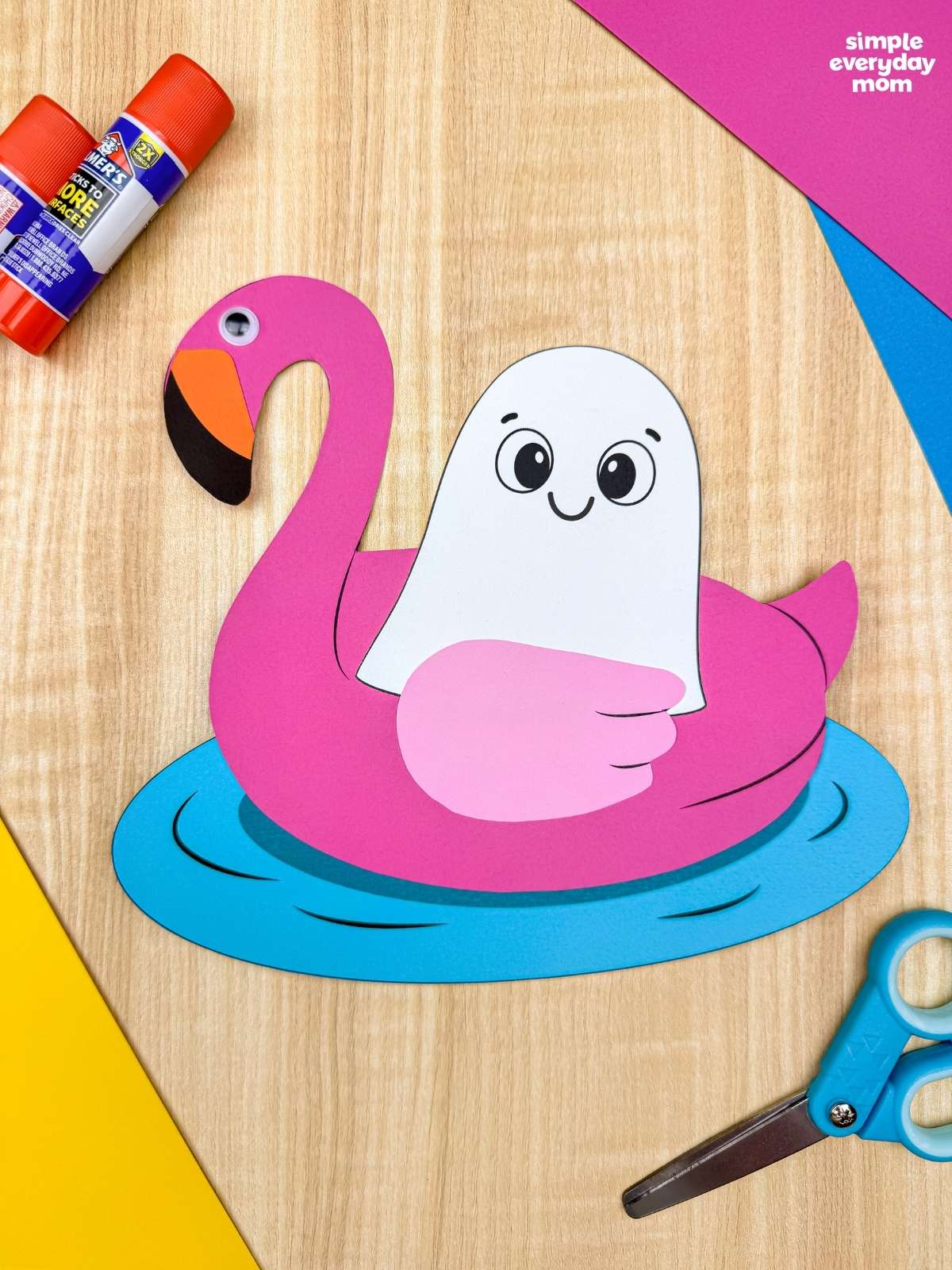 A completed craft featuring a Summerween ghost floating in a pink flamingo pool float surrounded by craft supplies.