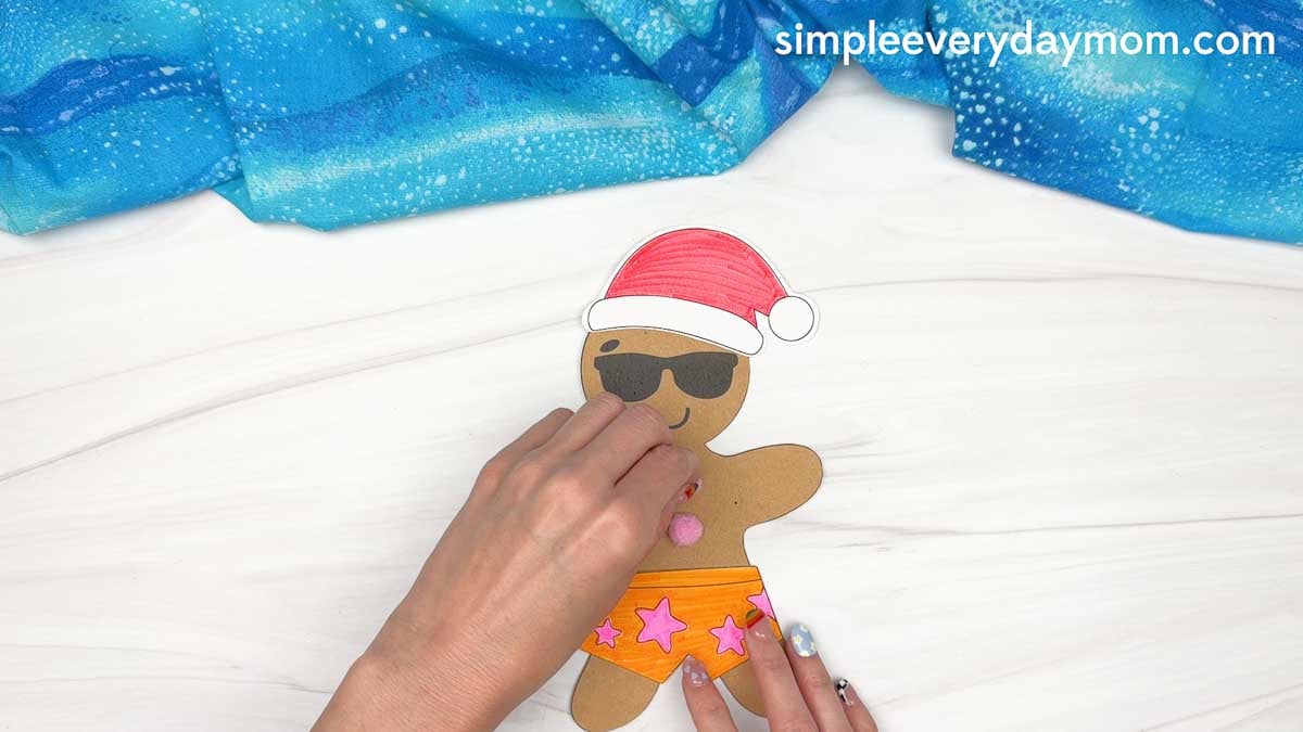 Hands glue a pink button onto a beach-themed gingerbread man wearing sunglasses, a Santa hat, and orange swim shorts for a Christmas in July craft.