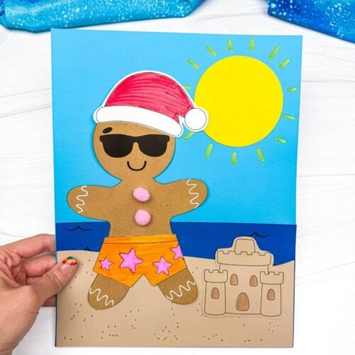 Hands hold a completed Christmas in July gingerbread beach craft featuring a gingerbread man in orange star swim shorts, sunglasses, a Santa hat, and a beach scene with a sandcastle and bright sun.