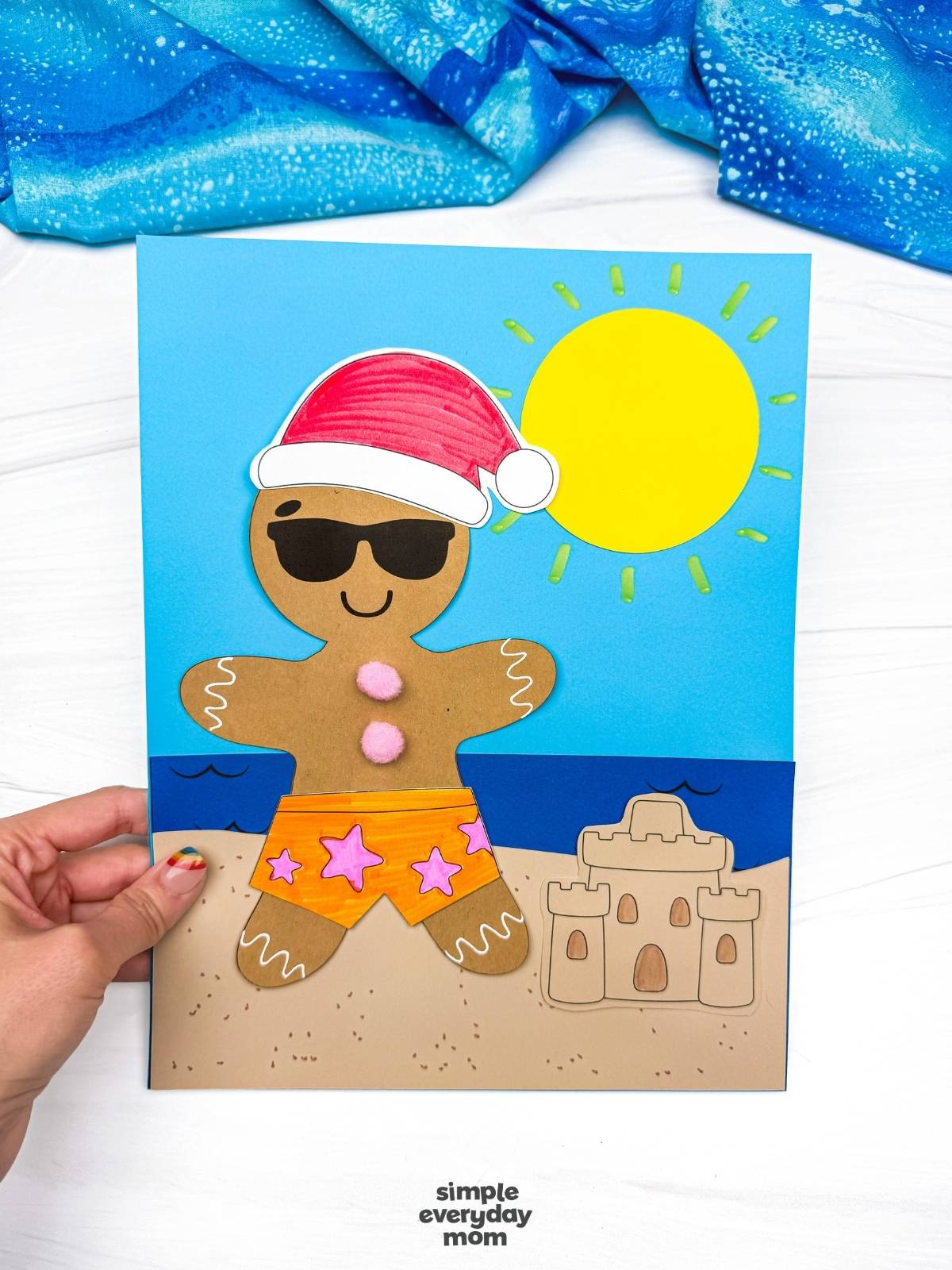 Hands hold a completed Christmas in July gingerbread beach craft featuring a gingerbread man in orange star swim shorts, sunglasses, a Santa hat, and a beach scene with a sandcastle and bright sun.