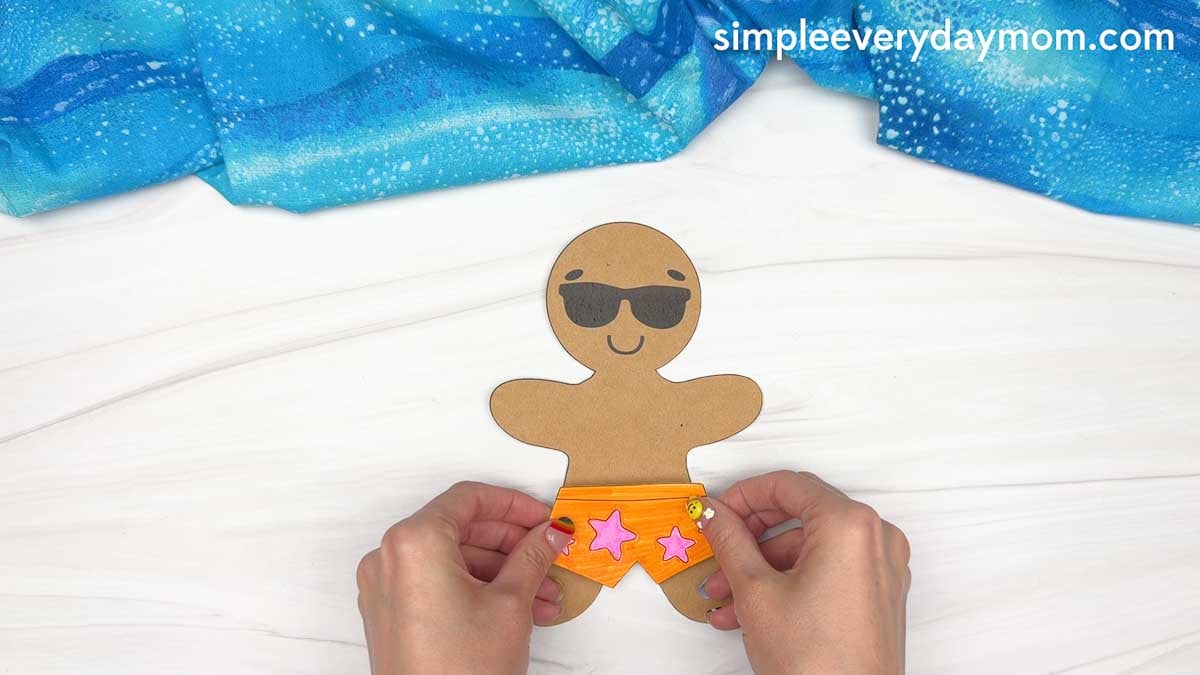 Hands attach orange beach shorts with pink star decorations to a sunglasses-wearing gingerbread man for a Christmas in July craft.