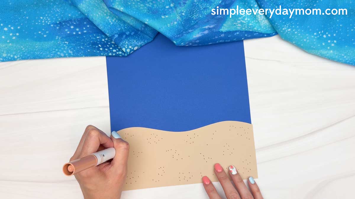 Hands drawing small dots on a tan paper beach piece attached to blue cardstock for a whale shark craft background.