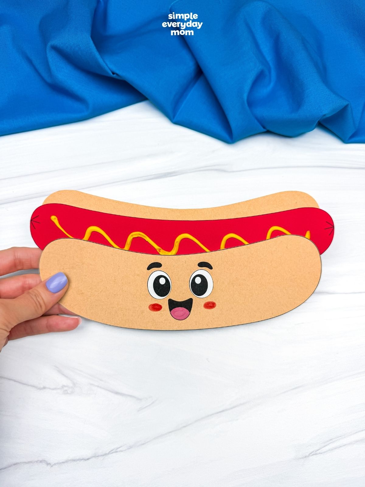 Hands holding a completed paper hot dog craft with a smiling face and mustard topping.