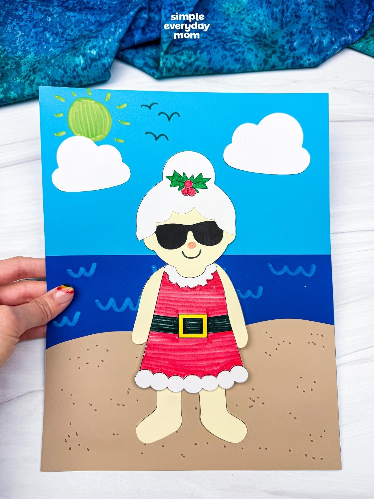 Hands holding a completed Summer Mrs. Claus craft on a sunny beach scene background.