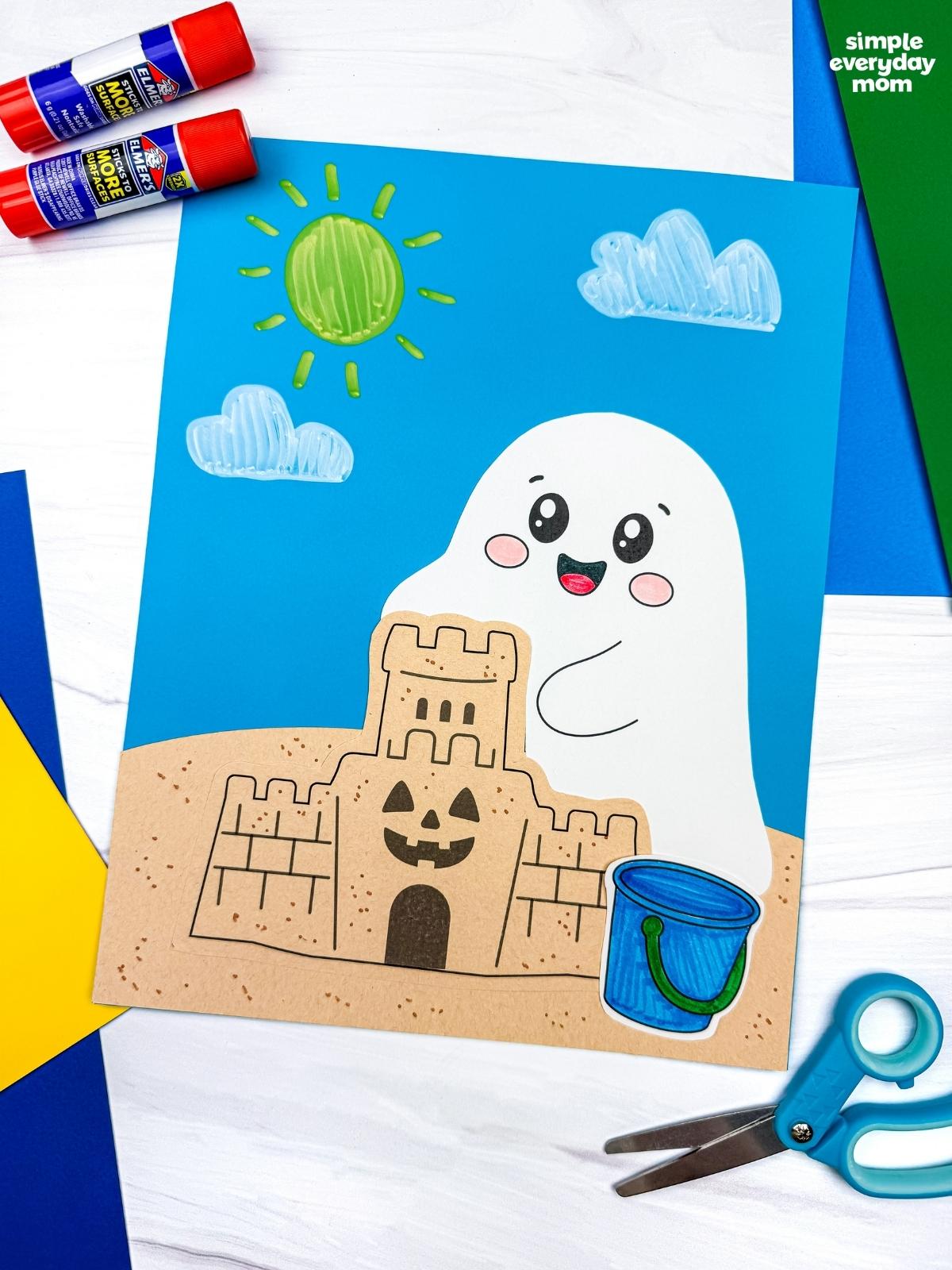 A completed Craft featuring a smiling beach ghost behind a sandcastle with a blue beach bucket, sunny sky, and fluffy clouds, surrounded by craft supplies.