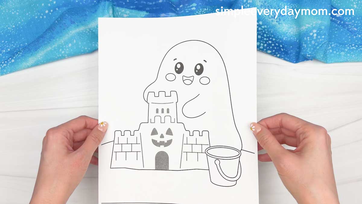 Hands holding a printable beach ghost craft template featuring a smiling ghost building a sandcastle with a bucket nearby.