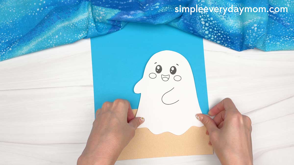 Hands placing a smiling ghost cutout onto the beach background for a beach ghost craft.