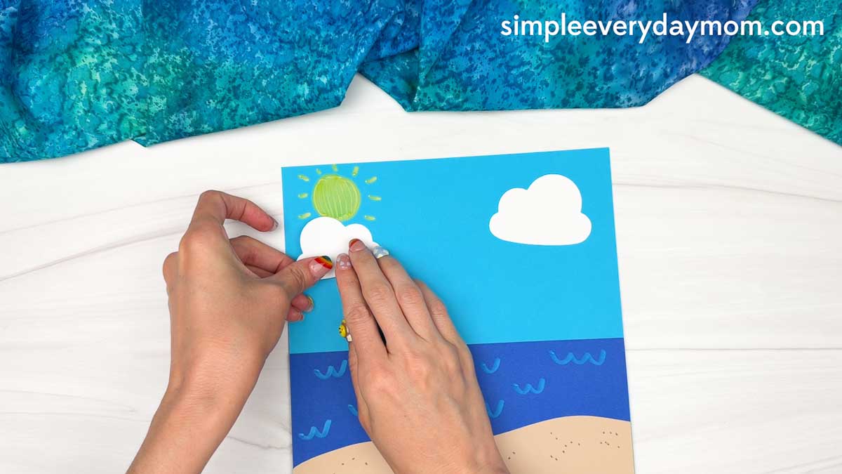 Hands placing a cloud cutout onto the beach scene background for the Summer Mrs. Claus craft.