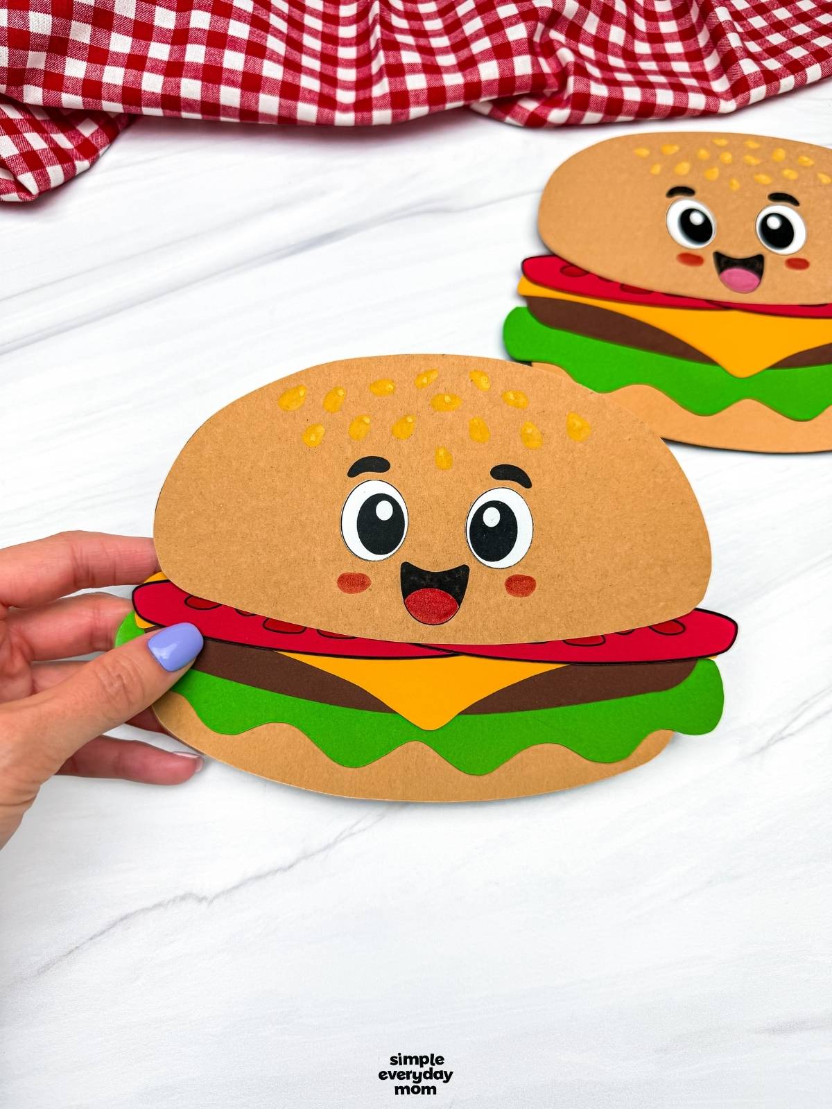 Hands holding a completed hamburger paper craft with a smiling cartoon face while another finished hamburger craft sits in the background.