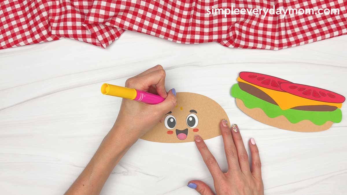Hands drawing sesame seeds on the cartoon hamburger top bun while the assembled burger layers sit nearby.