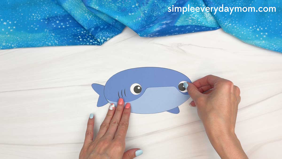 Hands attaching a googly eye to the whale shark craft template on a white tabletop.