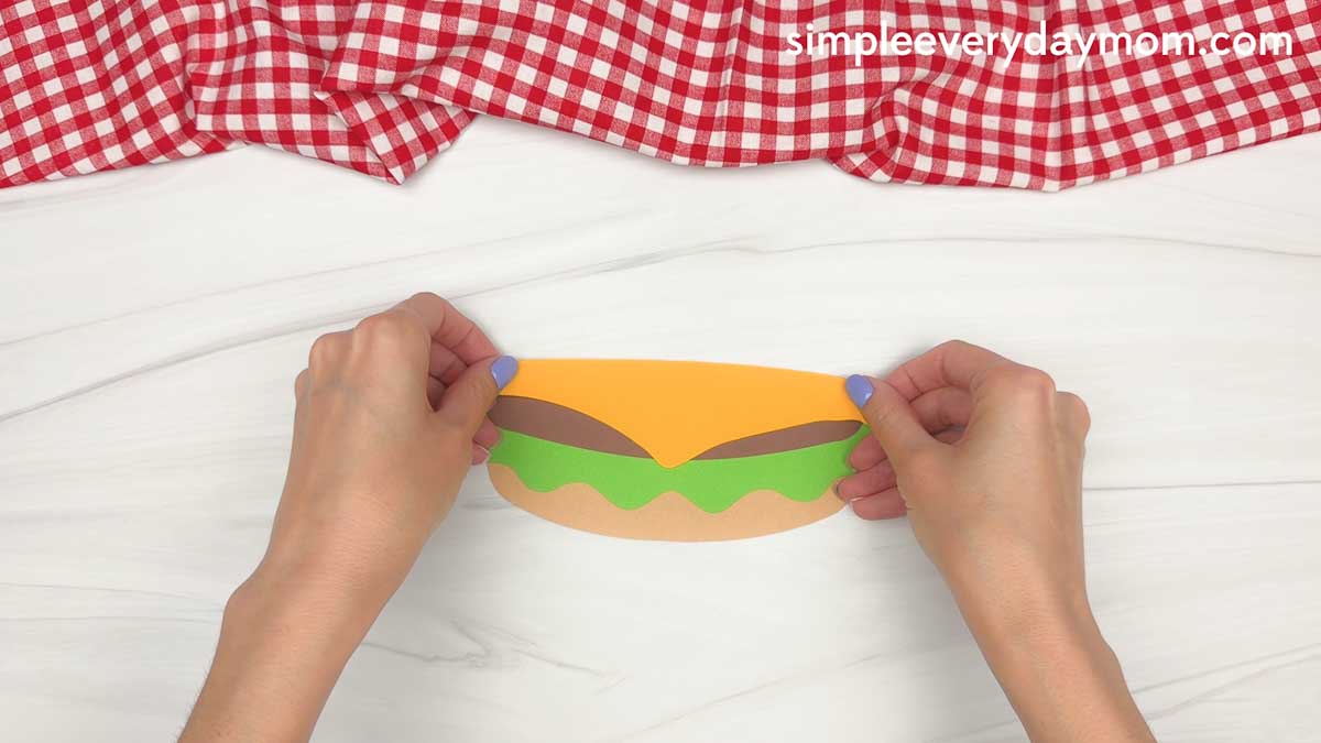 Hands adding a yellow paper cheese slice on top of the hamburger patty for a hamburger craft.