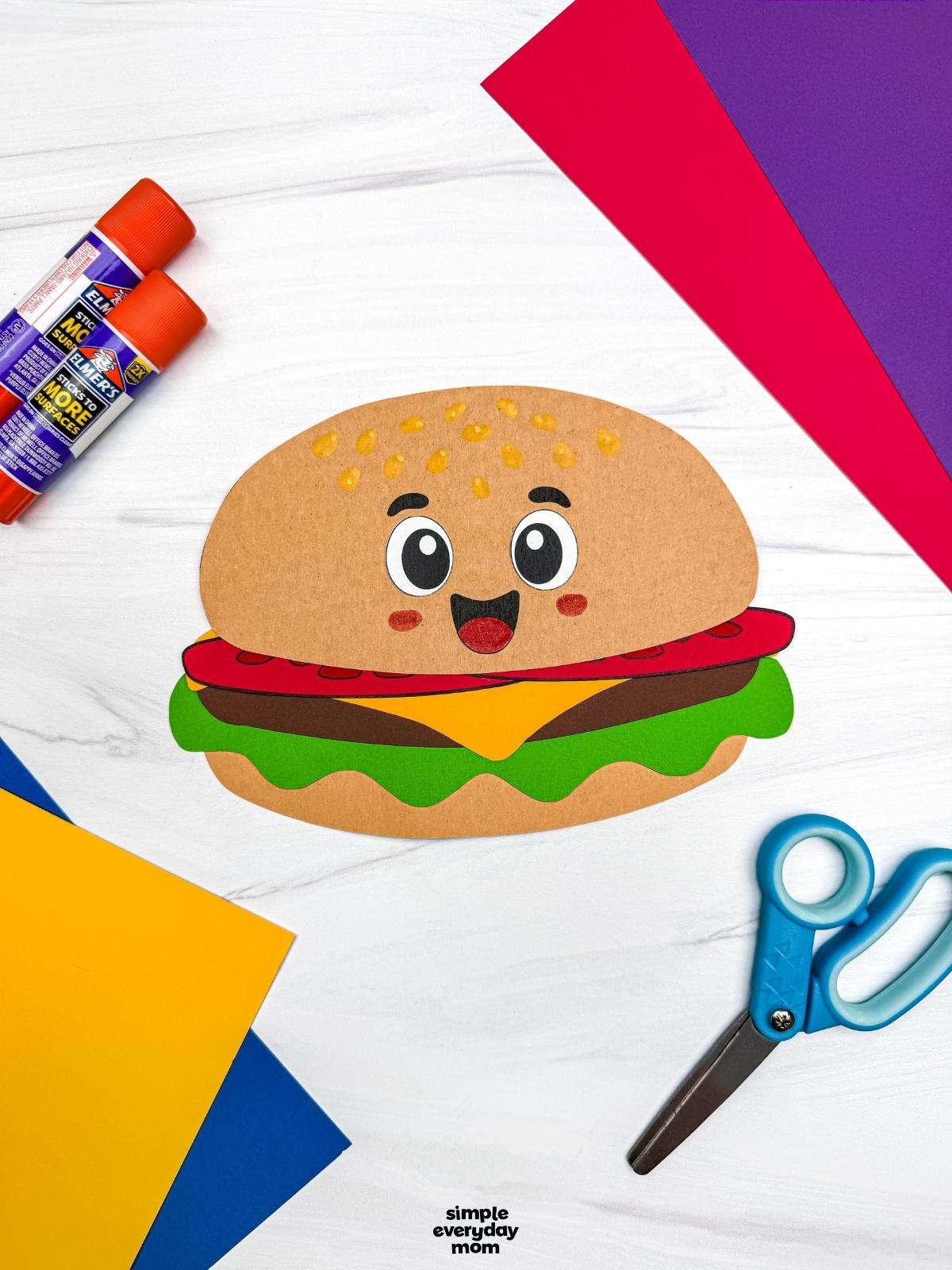 A completed hamburger paper craft with a smiling cartoon face displayed alongside scissors, glue sticks, and colorful cardstock.