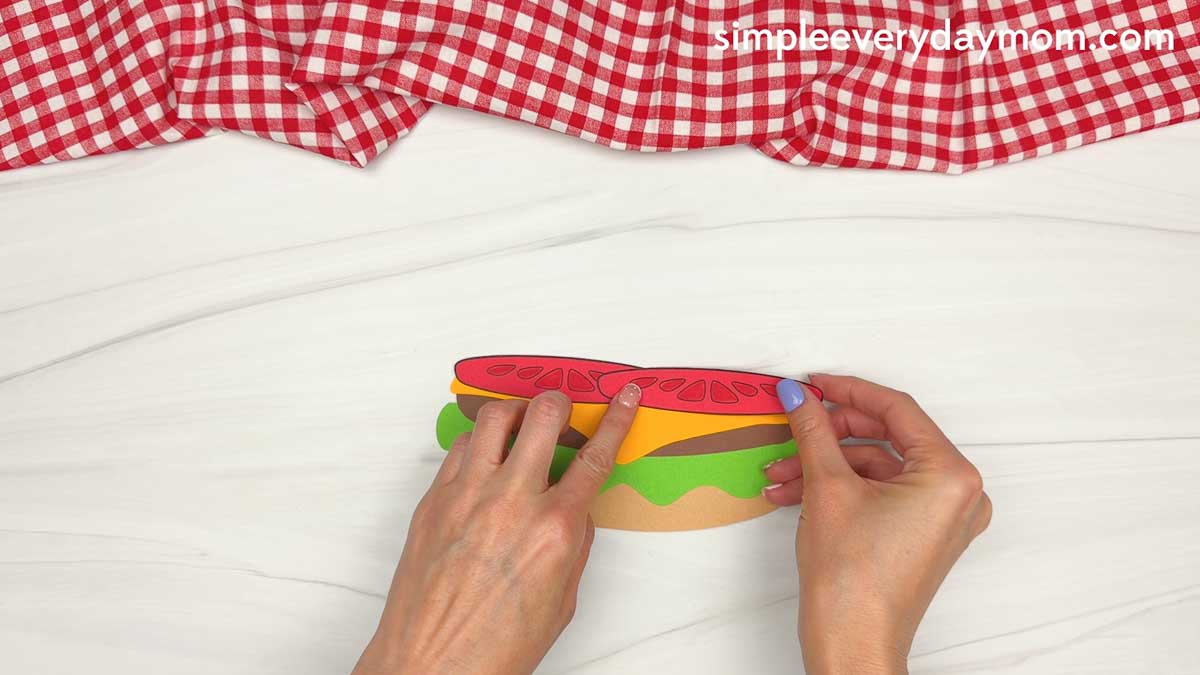Hands placing a red paper tomato slice on top of the cheese layer for a hamburger craft.
