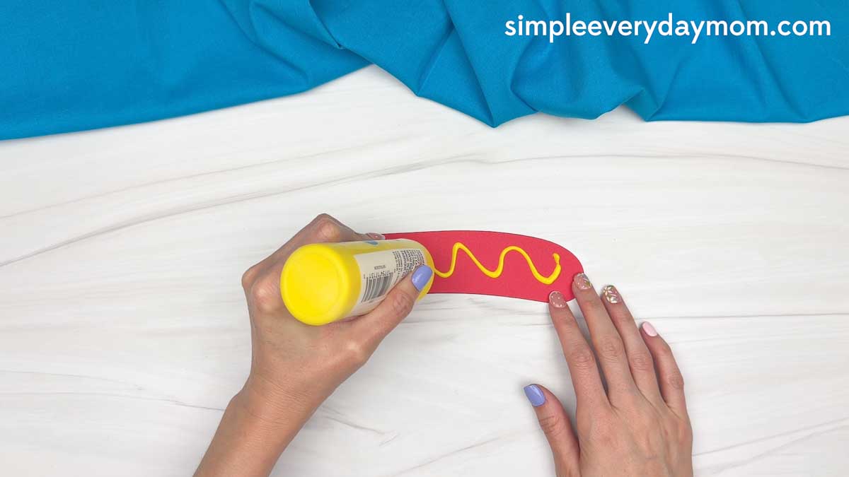 Hands adding a yellow squiggle of glue to a red paper hot dog cutout for a hot dog craft.