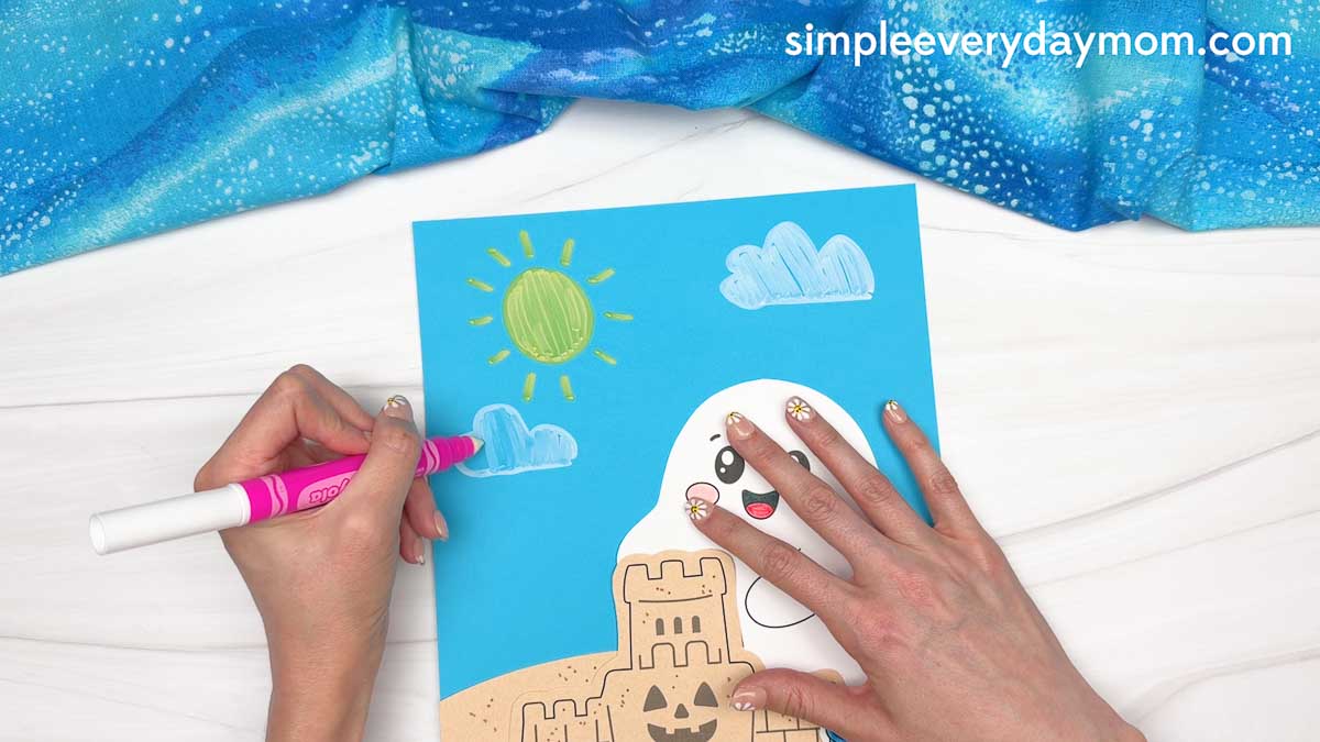 Hands drawing fluffy clouds above a smiling ghost and sandcastle on a beach ghost craft scene.