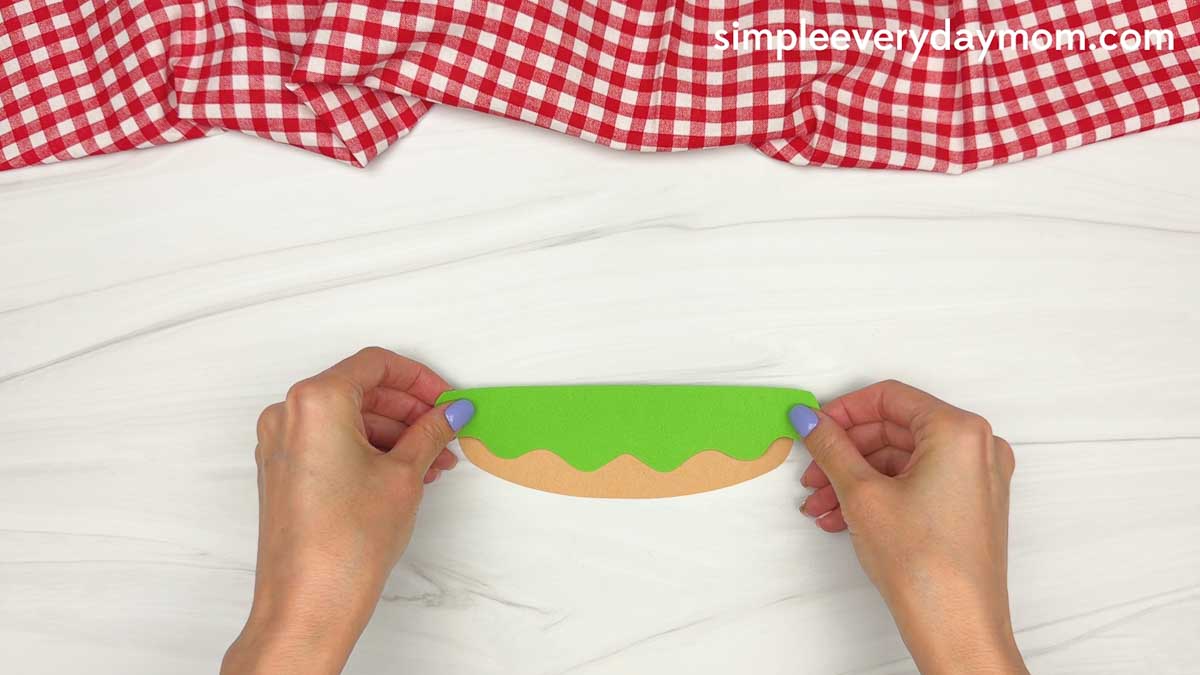 Hands placing a green paper lettuce piece onto the bottom bun for a hamburger craft.