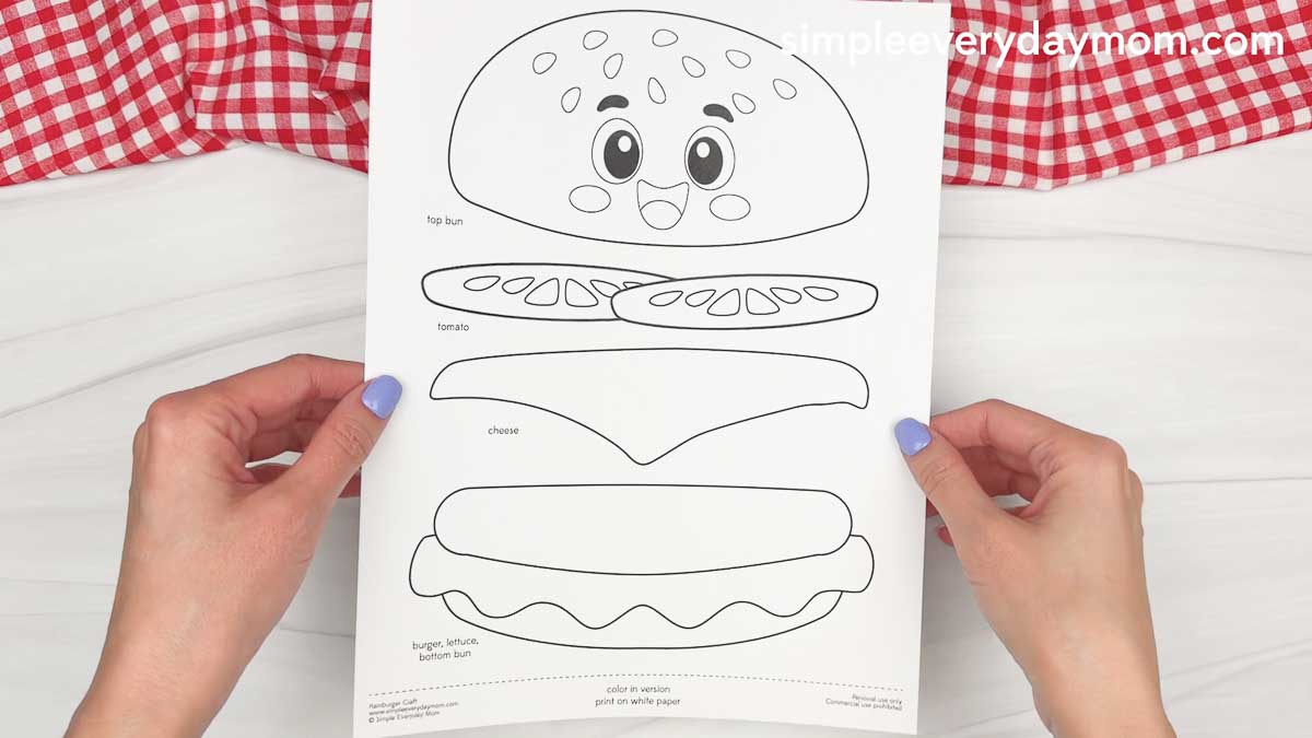 Hands holding a printed hamburger craft template with labeled burger pieces ready to color, cut, and assemble.