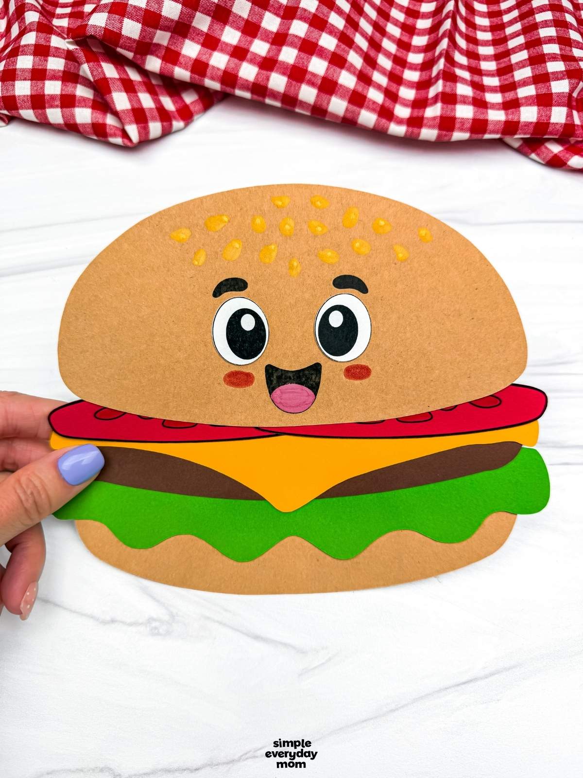 Hands holding a completed hamburger paper craft with a smiling cartoon face and colorful burger layers.