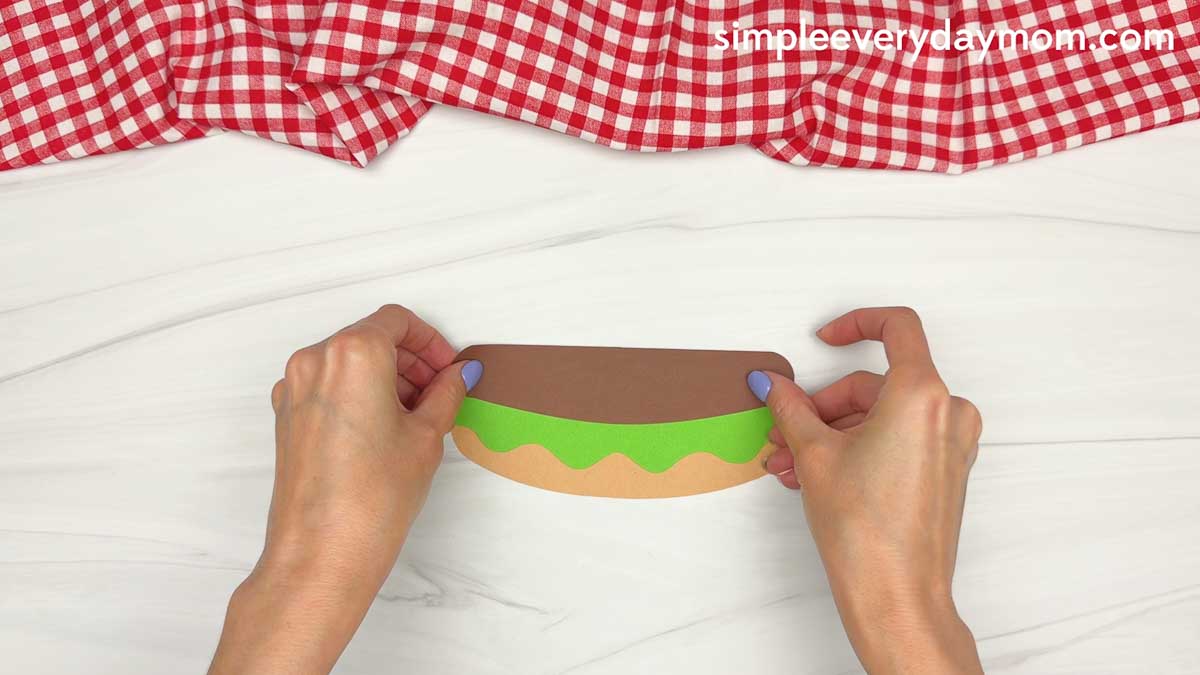 Hands placing a brown paper hamburger patty on top of the lettuce and bottom bun for a hamburger craft.