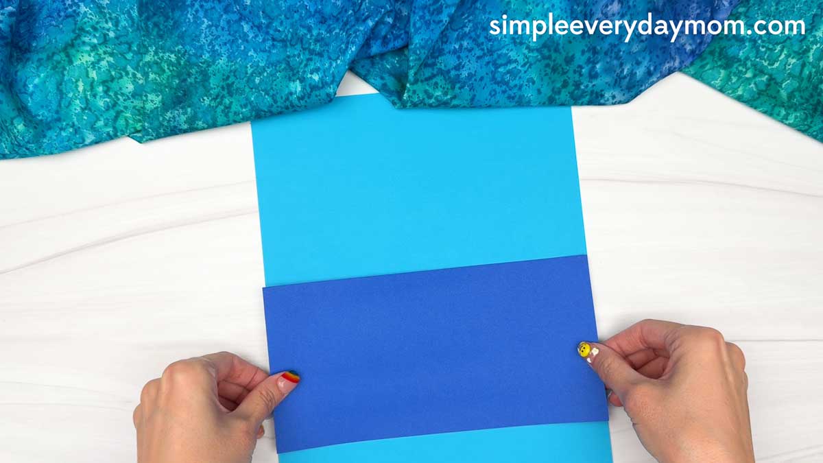 Hands positioning the ocean paper onto the beach scene background for the Summer Mrs. Claus craft.
