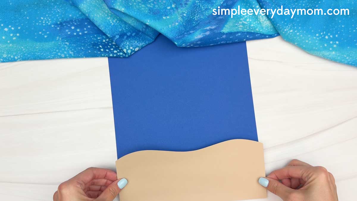 Hands positioning a tan paper beach piece onto blue cardstock to create the ocean background for a whale shark craft.