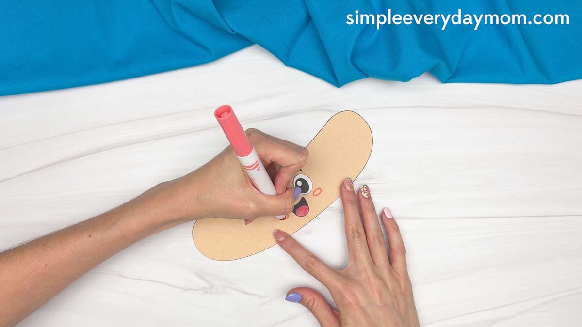 Hands drawing pink cheeks on a smiling paper hot dog bun cutout for a hot dog craft.