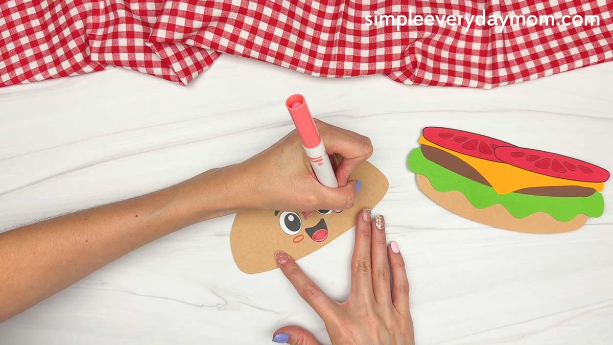 Hands coloring the cartoon hamburger face on the top bun while the assembled burger layers sit nearby.