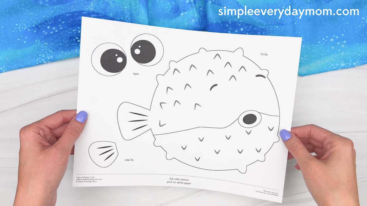 Hands holding a printed pufferfish paper craft template with cutout pieces for the body, eyes, and side fin.
