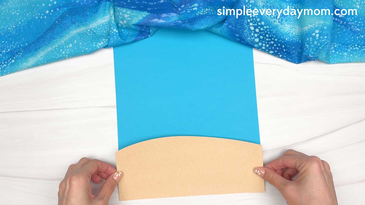 Hands positioning a tan sand cutout onto blue paper to create the beach background for a ghost craft.