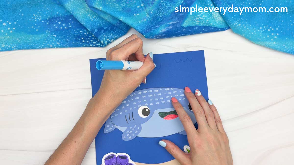 Hands drawing simple wave lines on the blue ocean background above a completed whale shark paper craft.