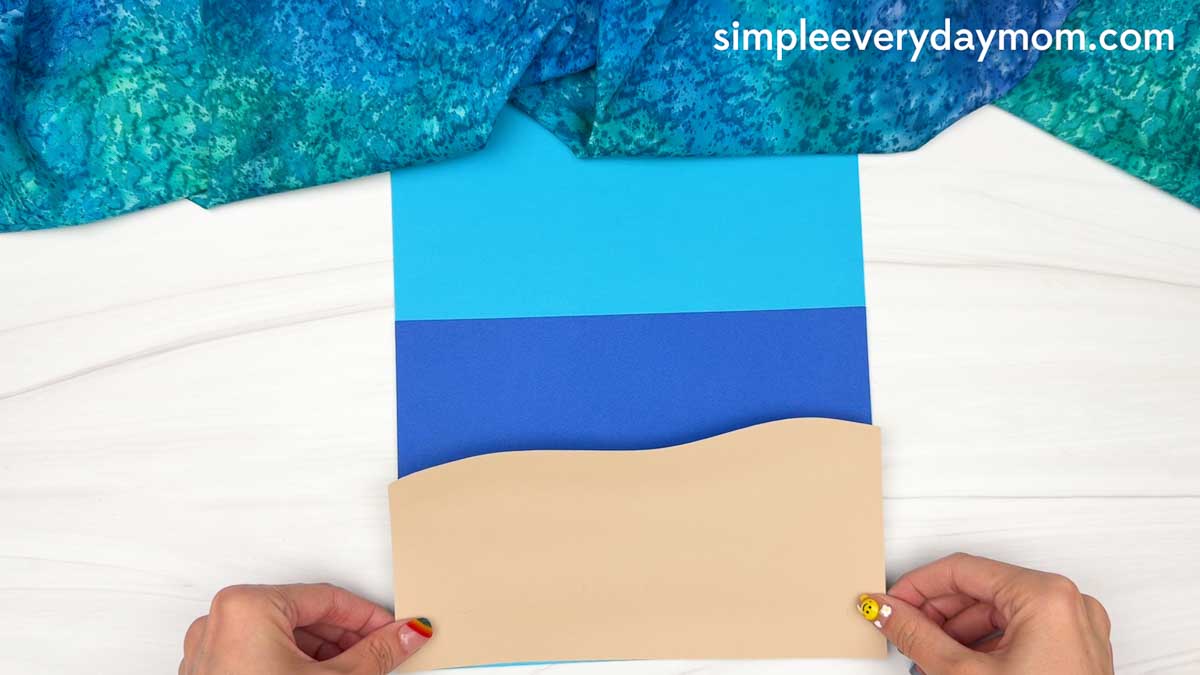 Hands positioning the sand paper onto the beach scene background for the Summer Mrs. Claus craft.