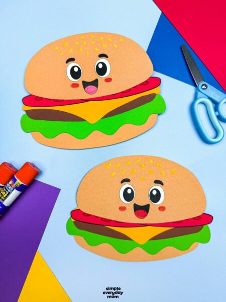A completed hamburger paper craft displayed with colorful cardstock, glue sticks, and scissors on a craft workspace.