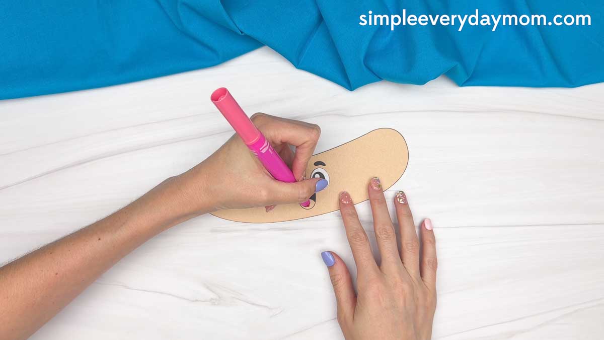 Hands coloring the tongue on a smiling paper hot dog bun cutout for a hot dog craft.