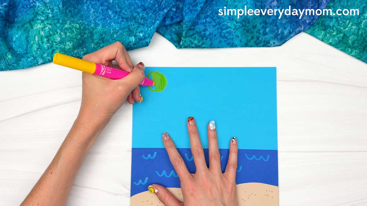 Hands drawing the sun on the beach scene background for the Summer Mrs. Claus craft.