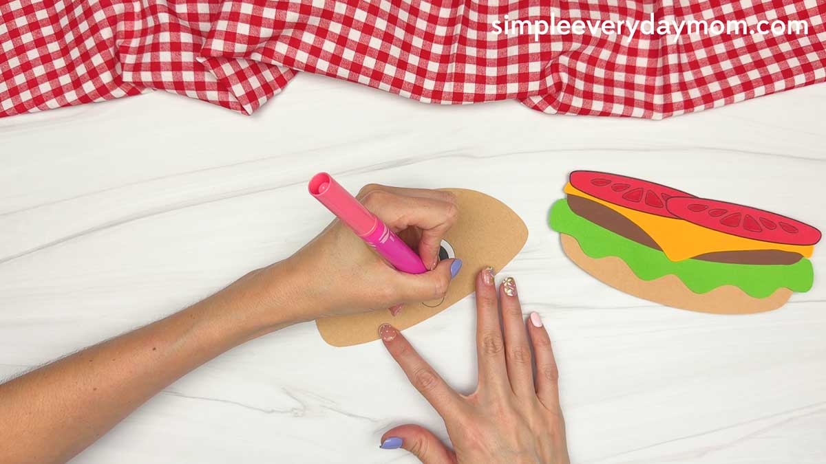 Hands drawing a smiling mouth on the hamburger top bun while the assembled burger layers rest beside it.