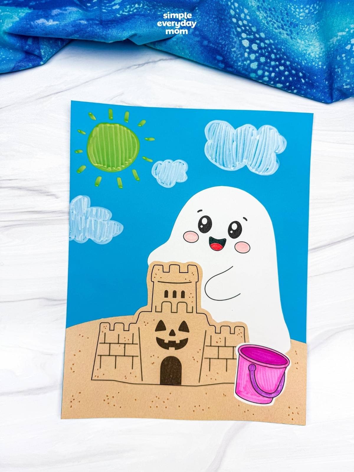 A completed Craft featuring a smiling beach ghost behind a sandcastle with a pink beach bucket, sunny sky, and fluffy clo