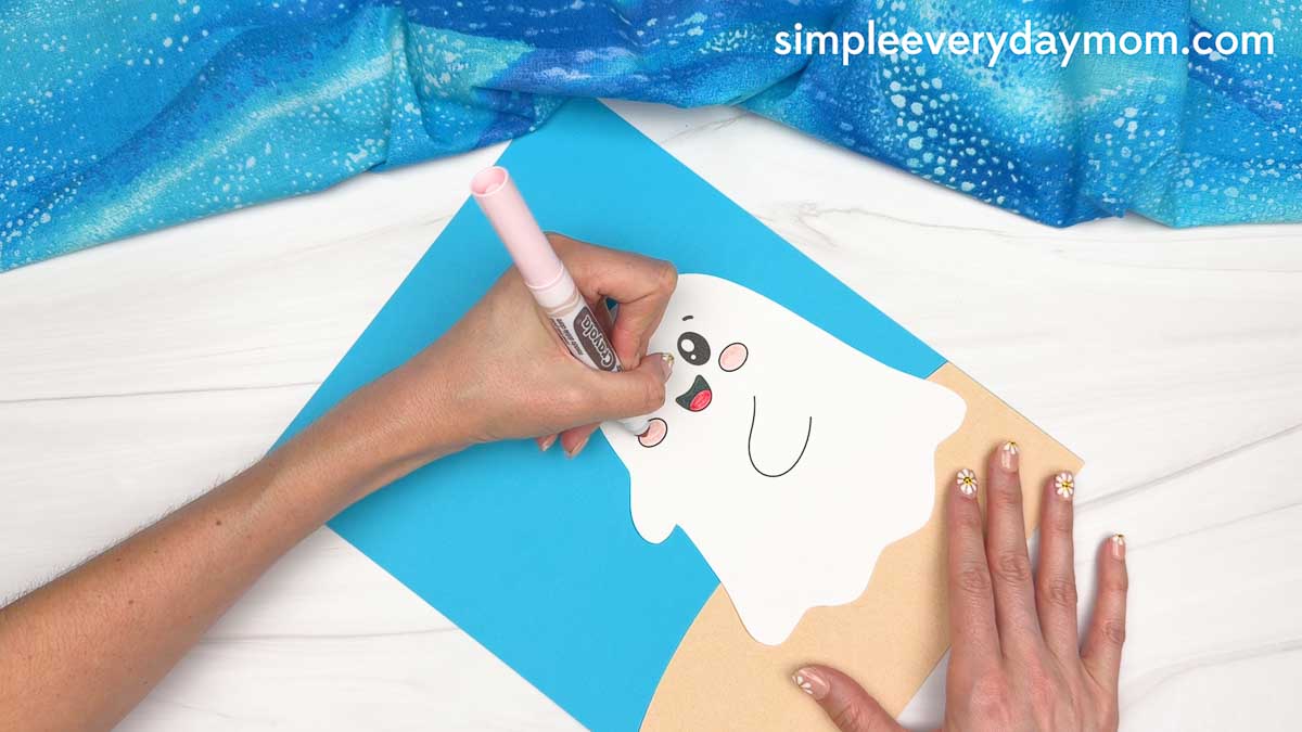 Hands coloring the ghost's cheeks pink while assembling a beach ghost craft on a sandy beach background.