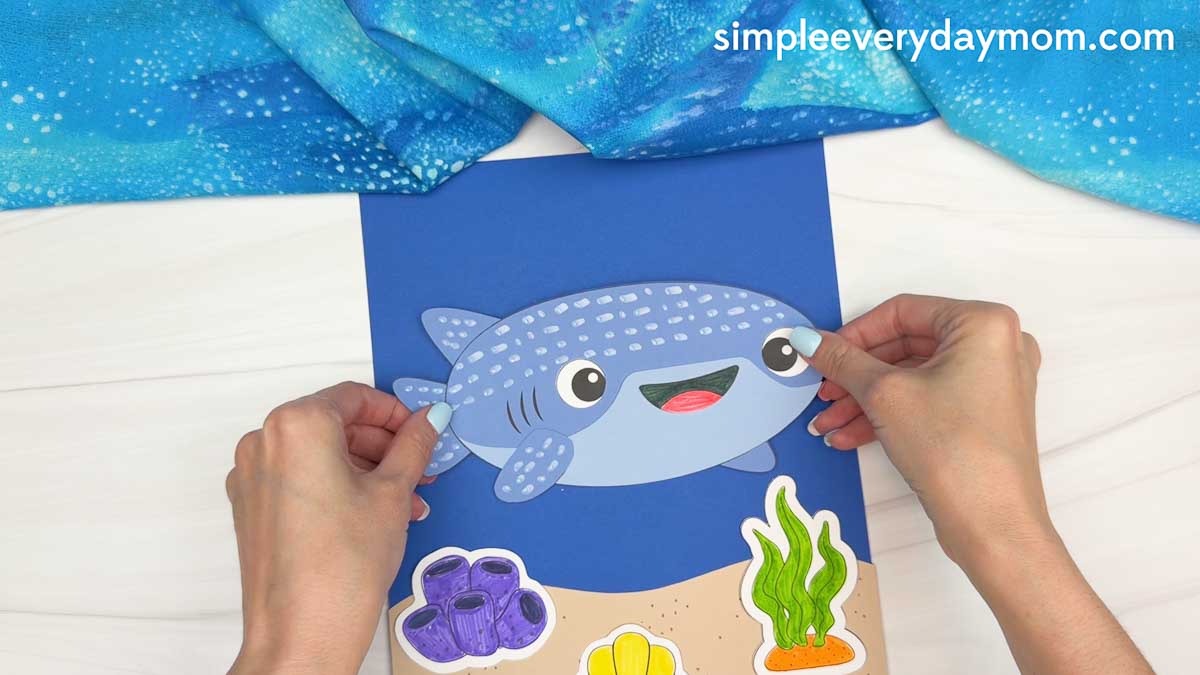 Hands placing the completed whale shark craft onto an ocean themed background with sand, seaweed, and coral decorations.