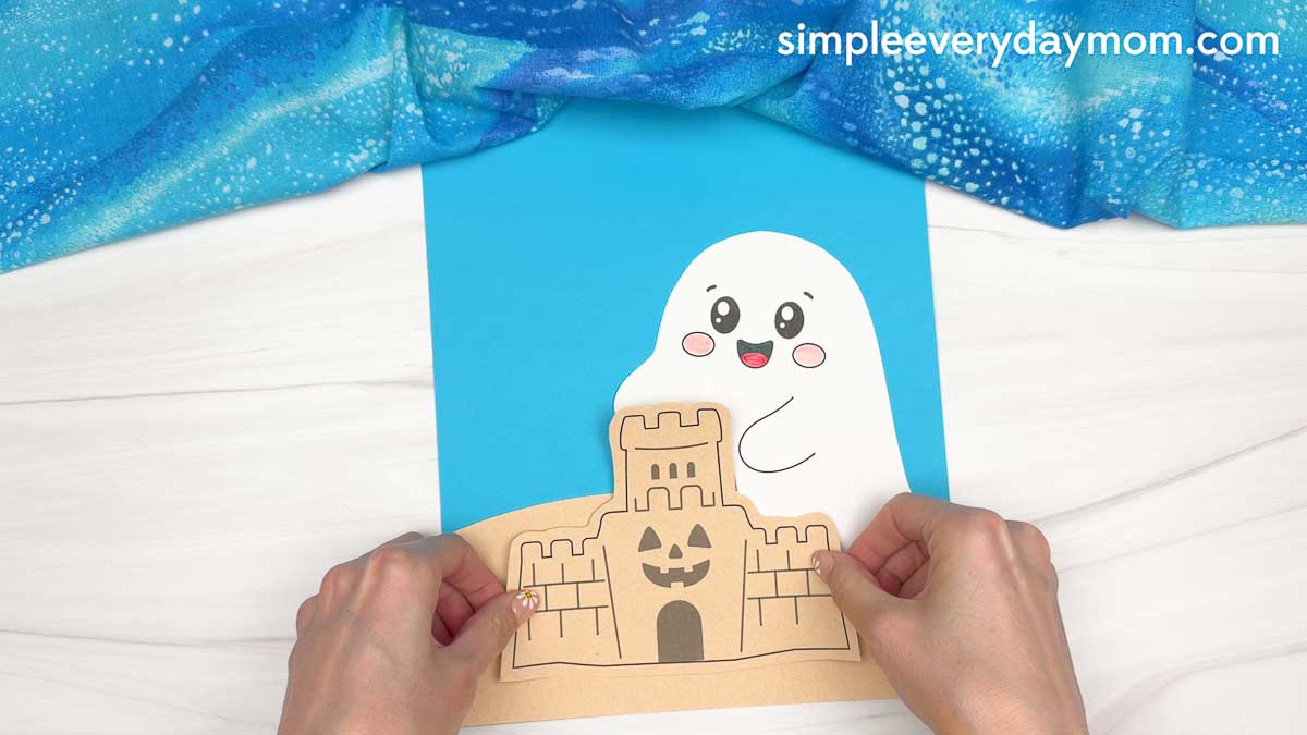 Hands placing a sandcastle cutout in front of a smiling ghost on the beach ghost craft background.