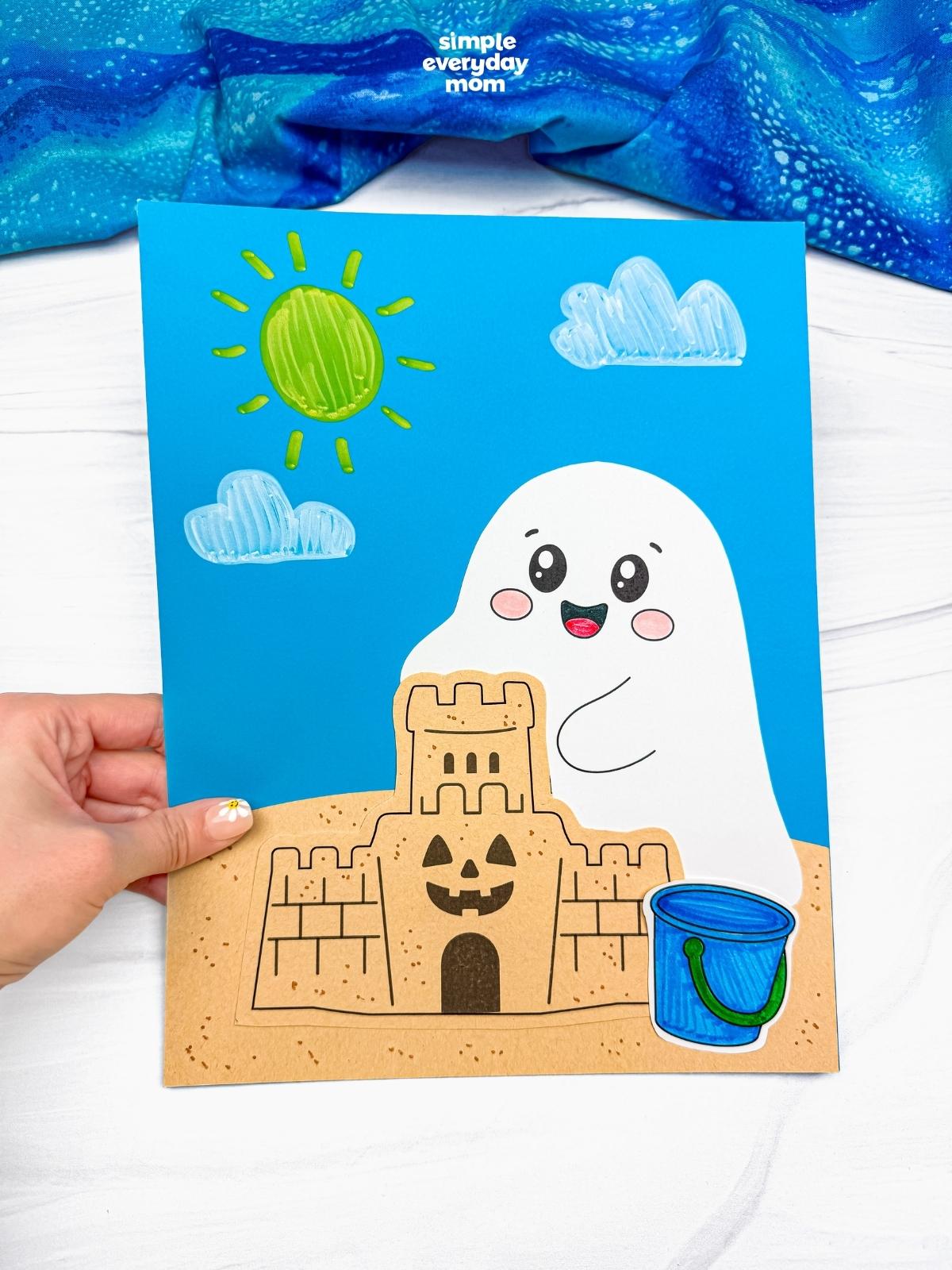 Hands holding a completed beach ghost craft featuring a smiling ghost, sandcastle, blue beach bucket, sunny sky, and fluffy clouds.