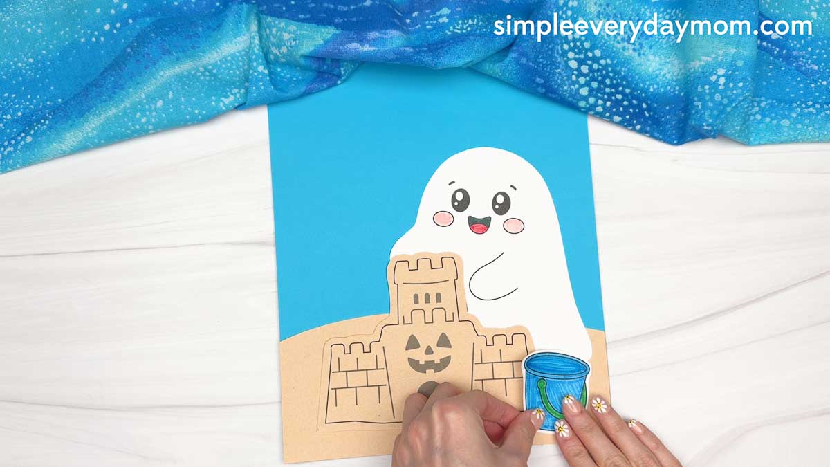 Hands attaching a blue beach bucket cutout beside the sandcastle on a beach ghost craft scene.